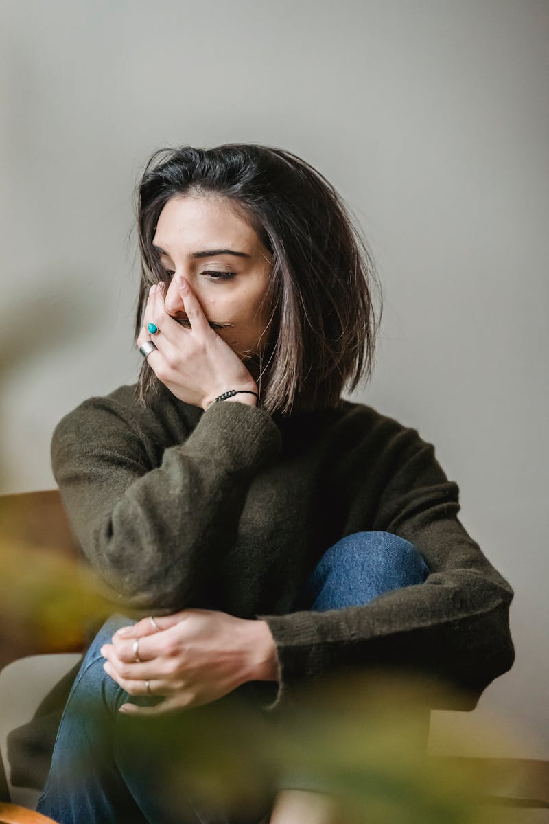 Unhappy female with dark hair in casual wear touching face while sitting in light room near white wall at home