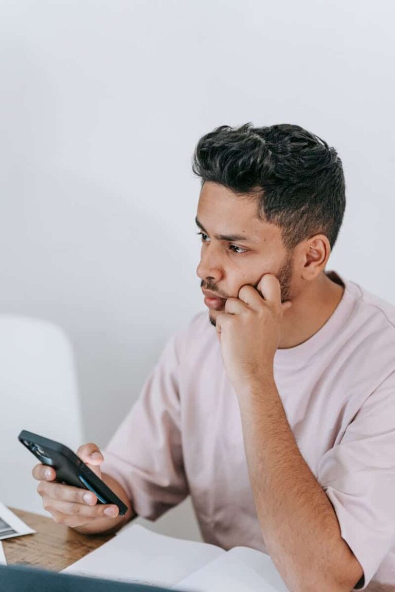 Pondering ethnic male in t shirt surfing mobile phone and touching face while sitting at desk with laptop and looking away in contemplation