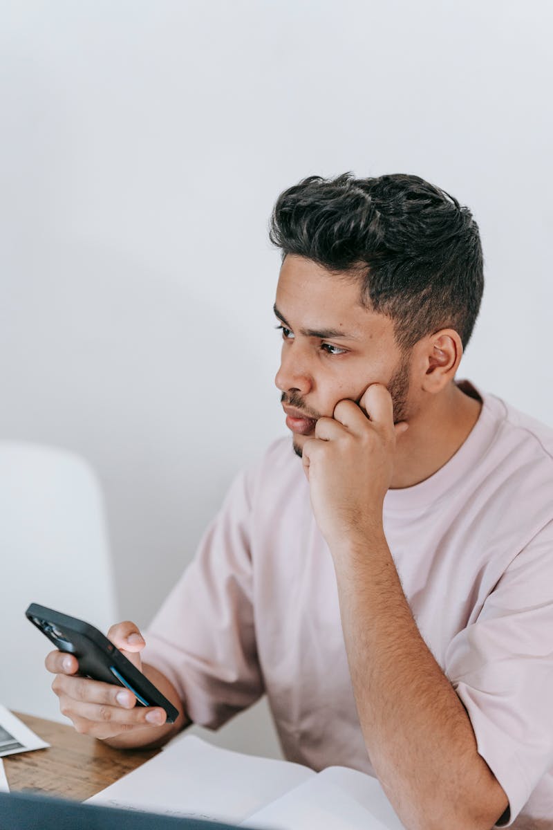 Pondering ethnic male in t shirt surfing mobile phone and touching face while sitting at desk with laptop and looking away in contemplation